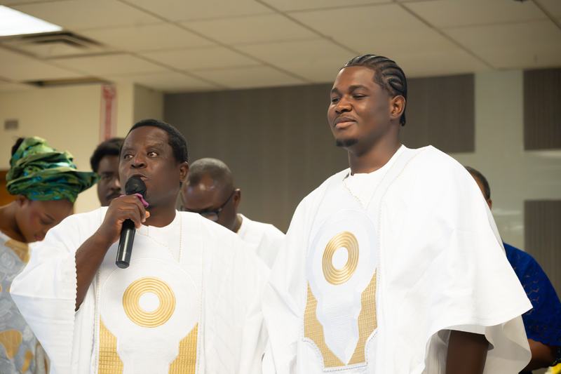 Two men in white agbada with gold embroidery, one speaking into a microphone